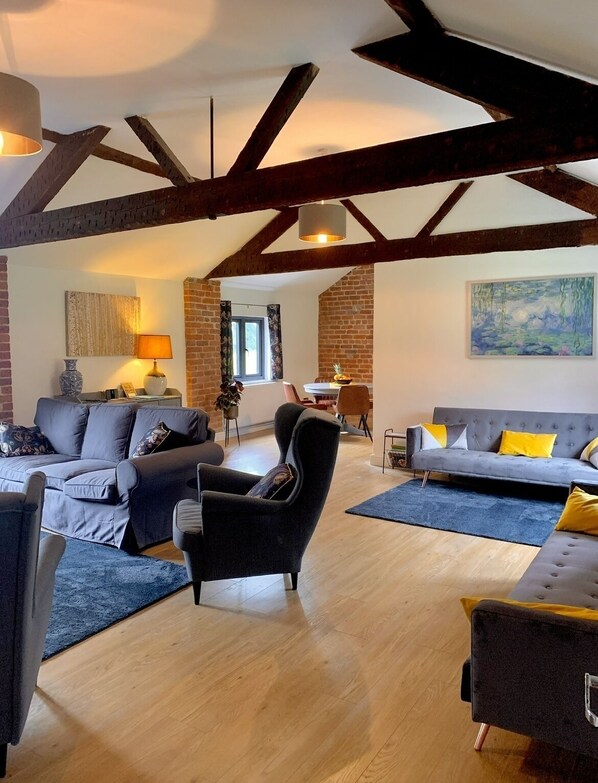 A spacious living room with exposed wooden beams, a grey sofa, a black armchair, and a blue rug.