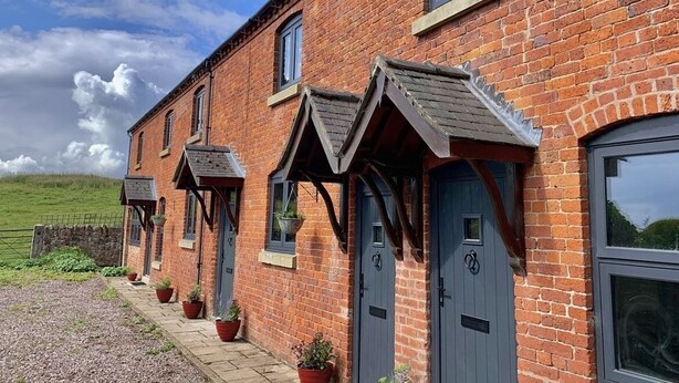 A row of brick houses with dark doors and windows, each with a small porch and potted plants. - New - Luxury Spacious Apartment in Beautiful Grounds (Oswestry, Shropshire)