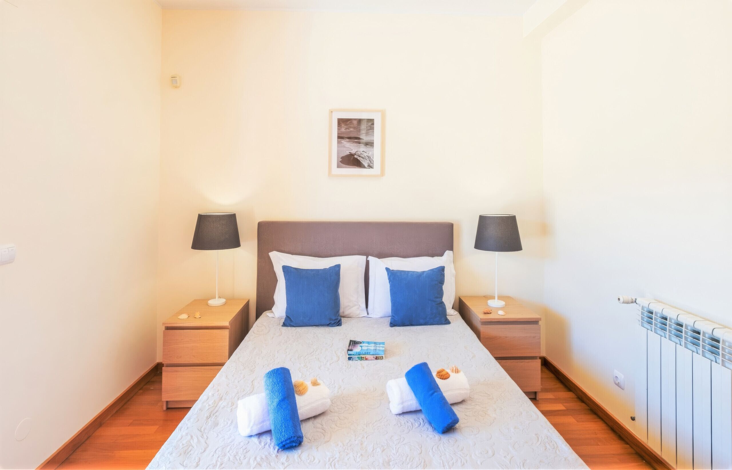 A hotel room with a bed, two bedside tables, a nightstand, a framed picture on the wall, and a radiator.