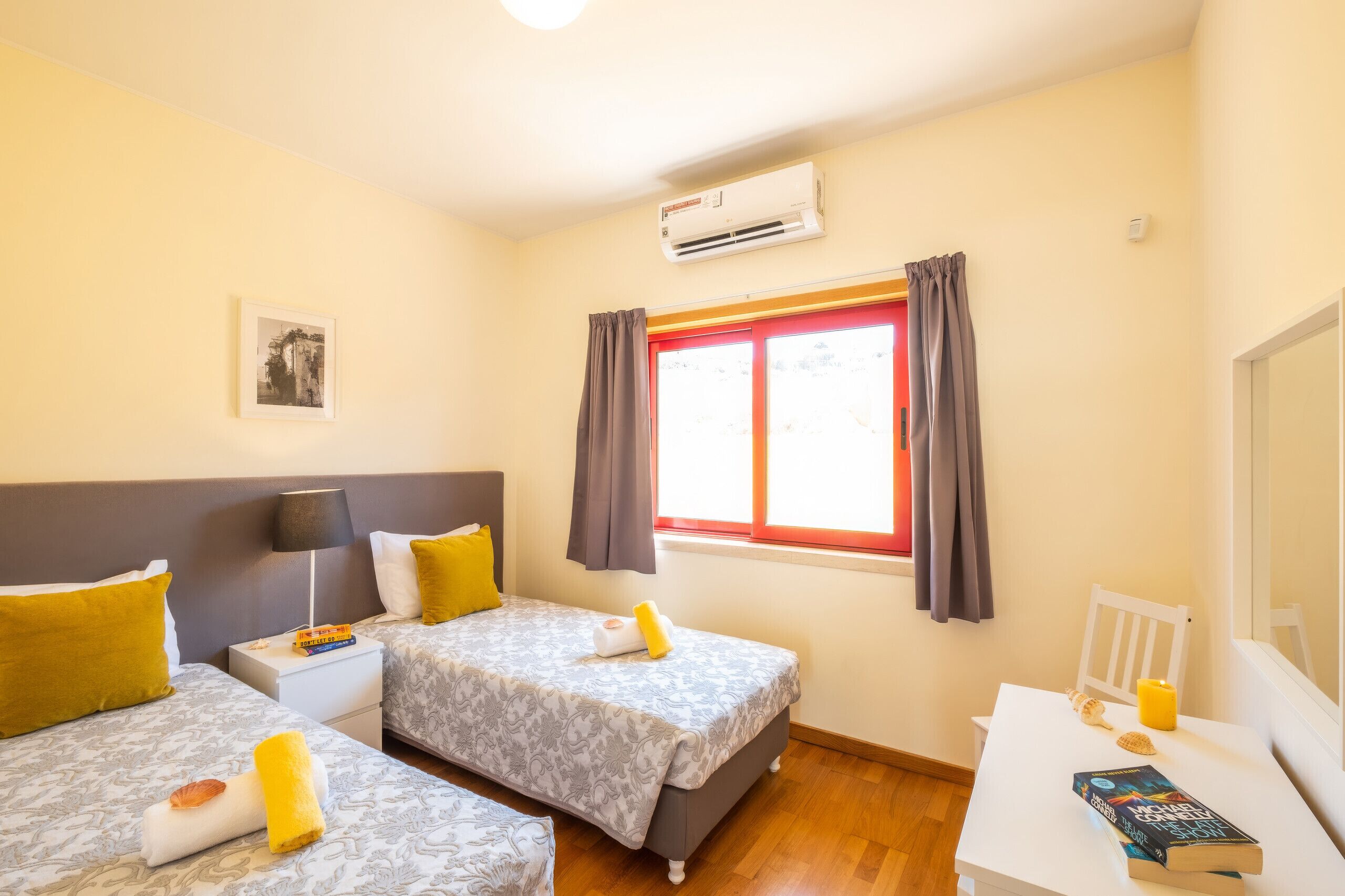 Habitación de hotel con dos camas, una mesita de noche con lámpara, una pequeña mesa con un libro y una ventana con cortinas.