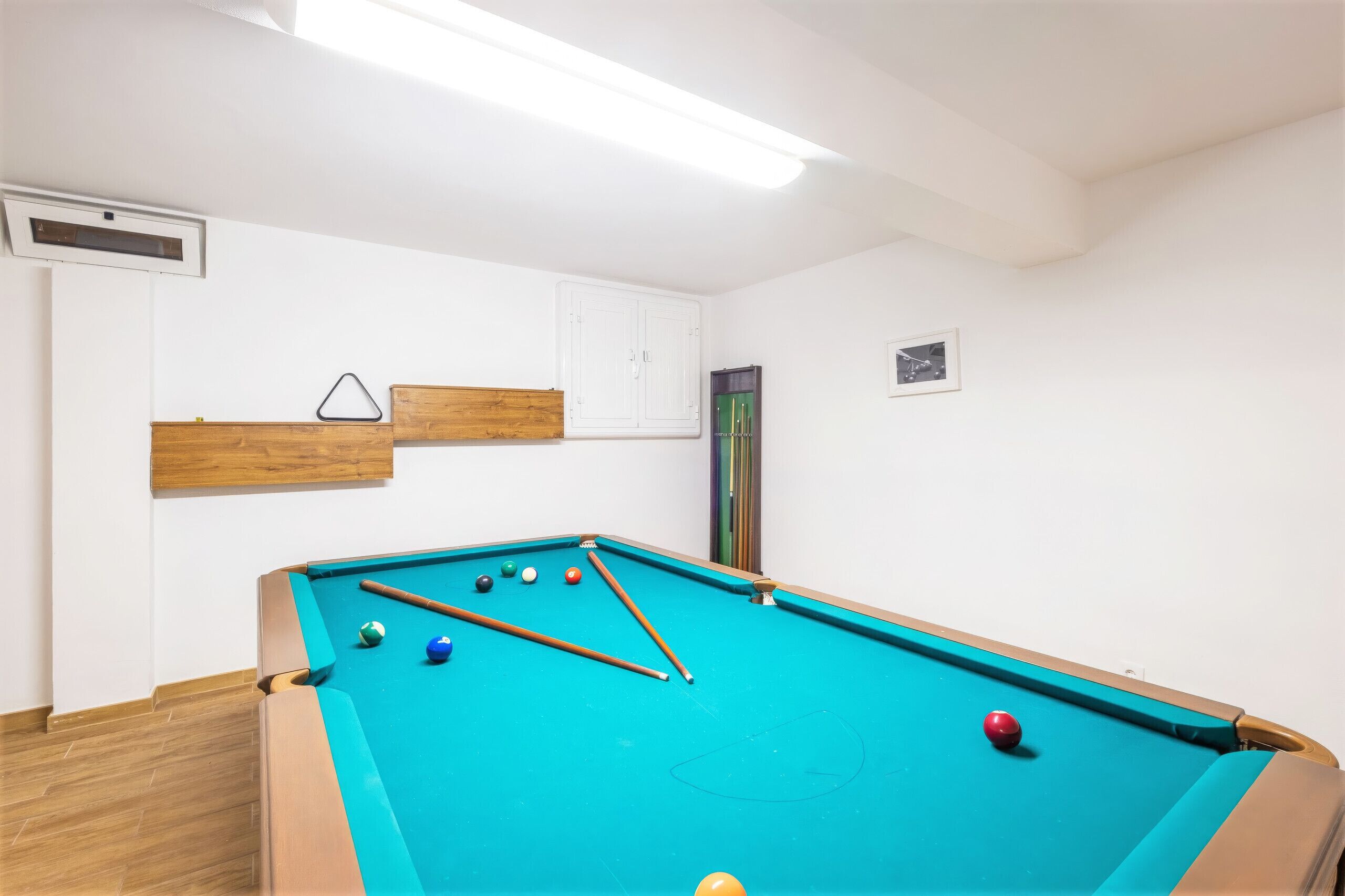 A pool table with balls and cues, a wooden bench, a white wall with a framed picture, and a door.