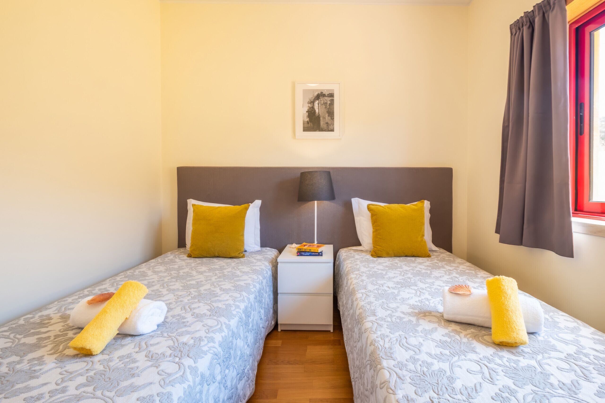 A double room with two beds, each with a patterned bedspread and a yellow pillow. There is a nightstand with a lamp and a book between the beds. A framed picture hangs on the wall above the beds.