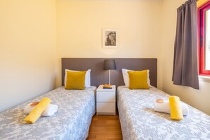 A double room with two beds, each with a patterned bedspread and a yellow pillow. There is a nightstand with a lamp and a book between the beds. A framed picture hangs on the wall above the beds. - Villa with heatable pool & BBQ in Albufeira Marina (Albufeira)