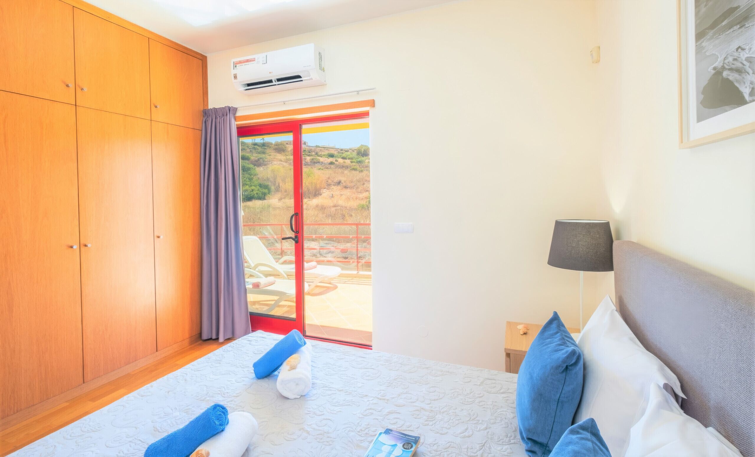 A hotel room with a bed, a nightstand, a lamp, a wardrobe, and a sliding glass door leading to an outdoor area.