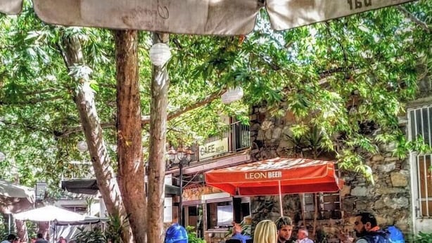 Ein Straßencafé mit Tischen und Stühlen, umgeben von Bäumen und Steinmauern.