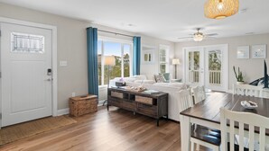 A modern living room with a sofa, dining table, and chairs. There is a ceiling fan and a hanging light fixture. - Marsh-View Oasis: 2-Min Easy Walk to Beach,Covered Decks, Outdoor Shower (Oak Island)