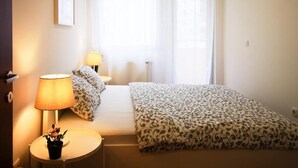 A hotel room with a bed, two bedside lamps, a small table with a plant, and a window with sheer curtains.