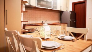 A kitchen with a wooden table set for two, white chairs, a refrigerator, and a clock on the wall.
