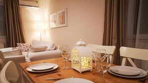 A dining area with a wooden table set for four, white plates, cutlery, and wine glasses. A decorative lantern is on the table, and a sofa is visible in the background.