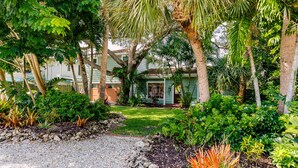 A lush garden with a variety of tropical plants and a gravel path leading to a house with a porch. - Designer Home on a Canal (Siesta Key)