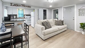 A modern living room with a sofa, coffee table, and chairs. There is a kitchen area with white cabinets and appliances. The room has a ceiling light and a door with a sign that reads "Lake House". - Pet Friendly Home by Lake Shawnee! (Topeka)