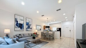 Una sala de estar moderna con un sofá gris, un comedor con mesa y sillas, y una cocina con refrigerador y estufa.