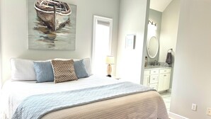 Un dormitorio con una cama grande, un cuadro de un barco, un baño con lavamanos y espejo, y una puerta que da a otra habitación.