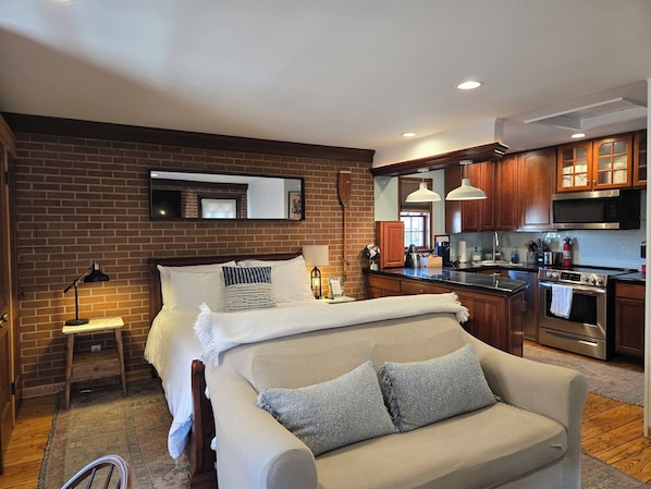 A compact living space with a kitchenette, a bed with pillows, a small table, and a lamp.
