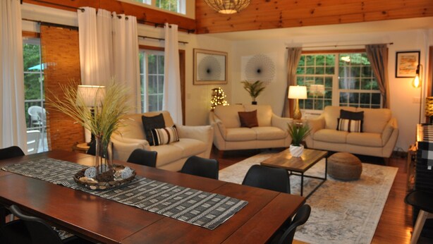 A cozy living room with two sofas, a dining table with a placemat, and a hanging light fixture. - Take me to the Trees! Lakefront on Autumn Lake, 3min to Salmon River (Orwell)