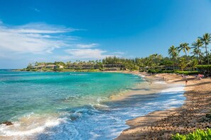 Une plage à l’eau turquoise et limpide, un rivage sablonneux et un complexe hôtelier comprenant plusieurs bâtiments et des palmiers.