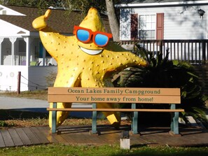 A large yellow starfish sculpture with sunglasses, a wooden bench, and a sign that reads 'Ocean Lakes Family Campground - Your home away from home!'