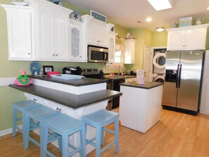 A kitchen with white cabinets, a stainless steel refrigerator, a microwave, and a sink. There are blue stools and a small island with a pink flamingo decoration.