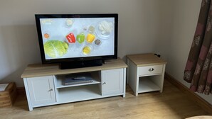 A television on a wooden TV stand with a remote control drawer, a small side table, and a decorative item on the floor. - Garden cottage self catering  (Collingham)