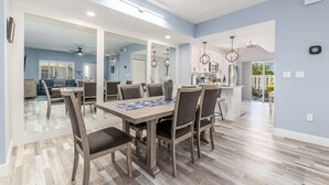 Un coin repas avec une table et des chaises en bois, une cuisine à l’arrière-plan et un ventilateur de plafond.