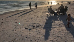 Une plage où l’on voit des gens se promener et s’asseoir sur le sable, et le soleil se couche à l’horizon.