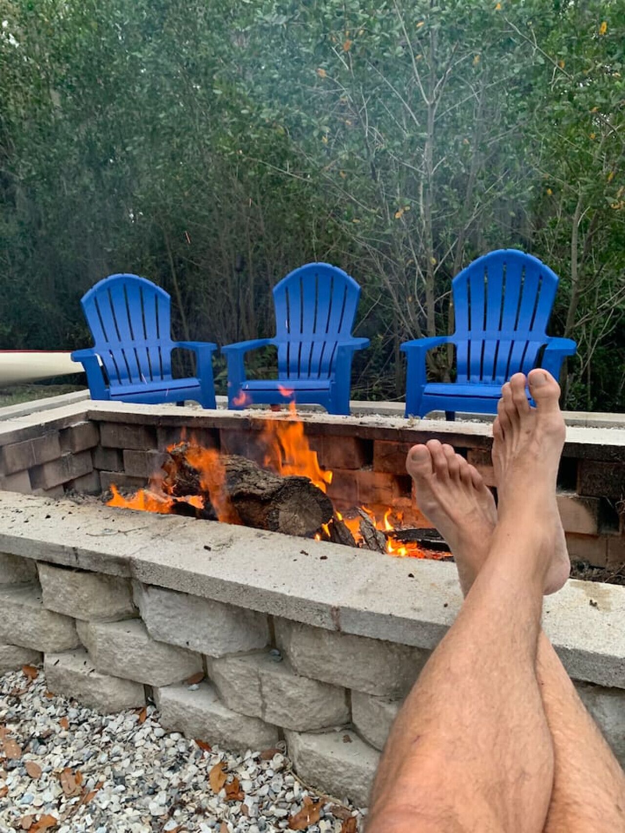 A fire pit with three blue Adirondack chairs and a person's legs resting on the edge.