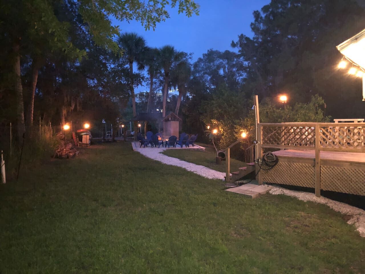 A well-lit outdoor patio area with a pathway leading to a seating area, surrounded by trees and a wooden fence.