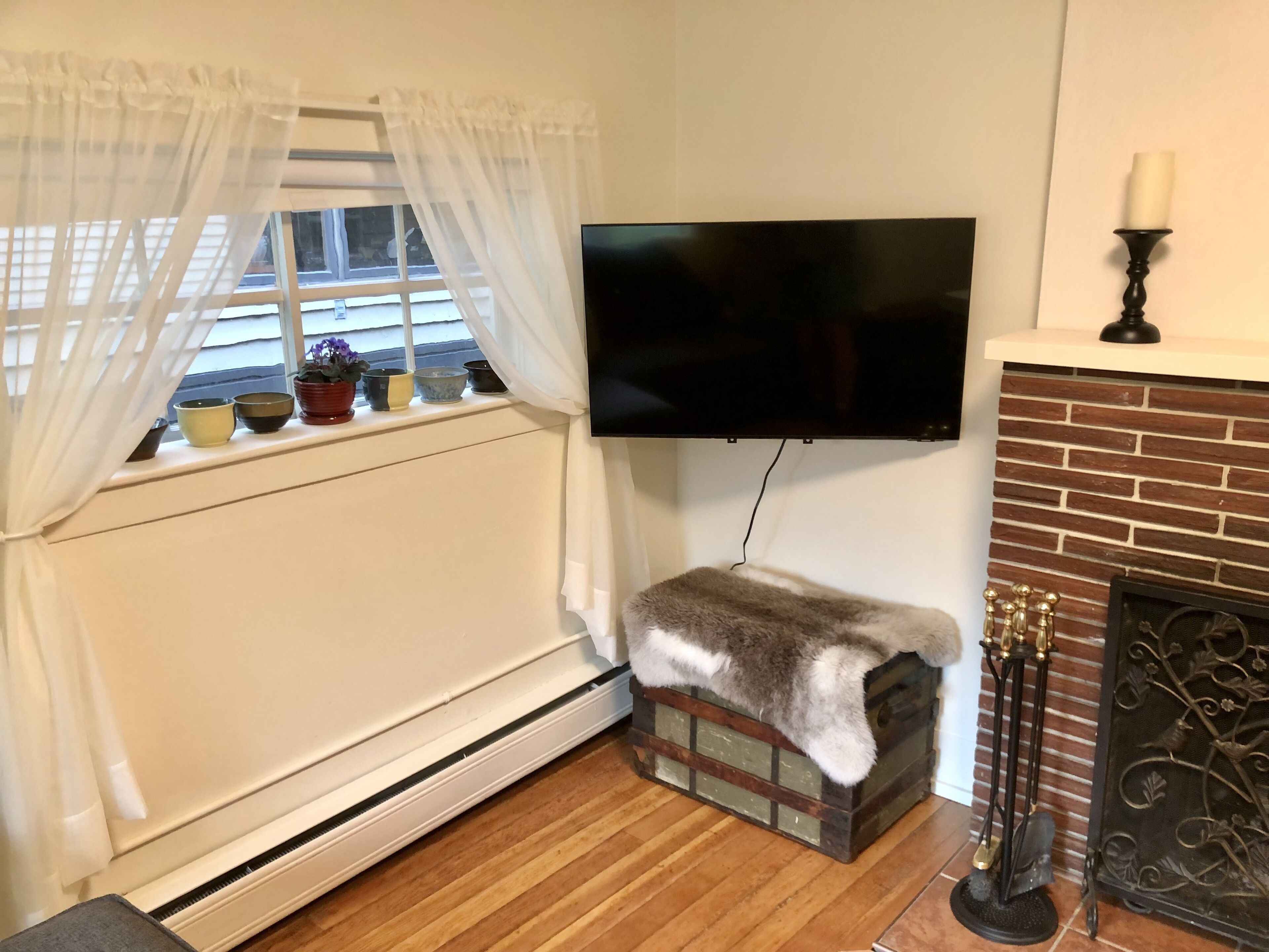 A room with a white radiator, a TV mounted on the wall, a fireplace with a mantel, and a window with sheer curtains.