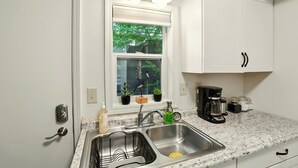 A kitchen with white cabinets, a double sink, a window with plants, and a coffee maker. - Coastal Cabin - Downtown Historical Log Home (Anchorage)