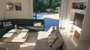Una sala de estar moderna con un sofá de cuero negro, una mesa de centro blanca y un sofá color beige. A la derecha hay una chimenea y se ve una piscina al exterior.