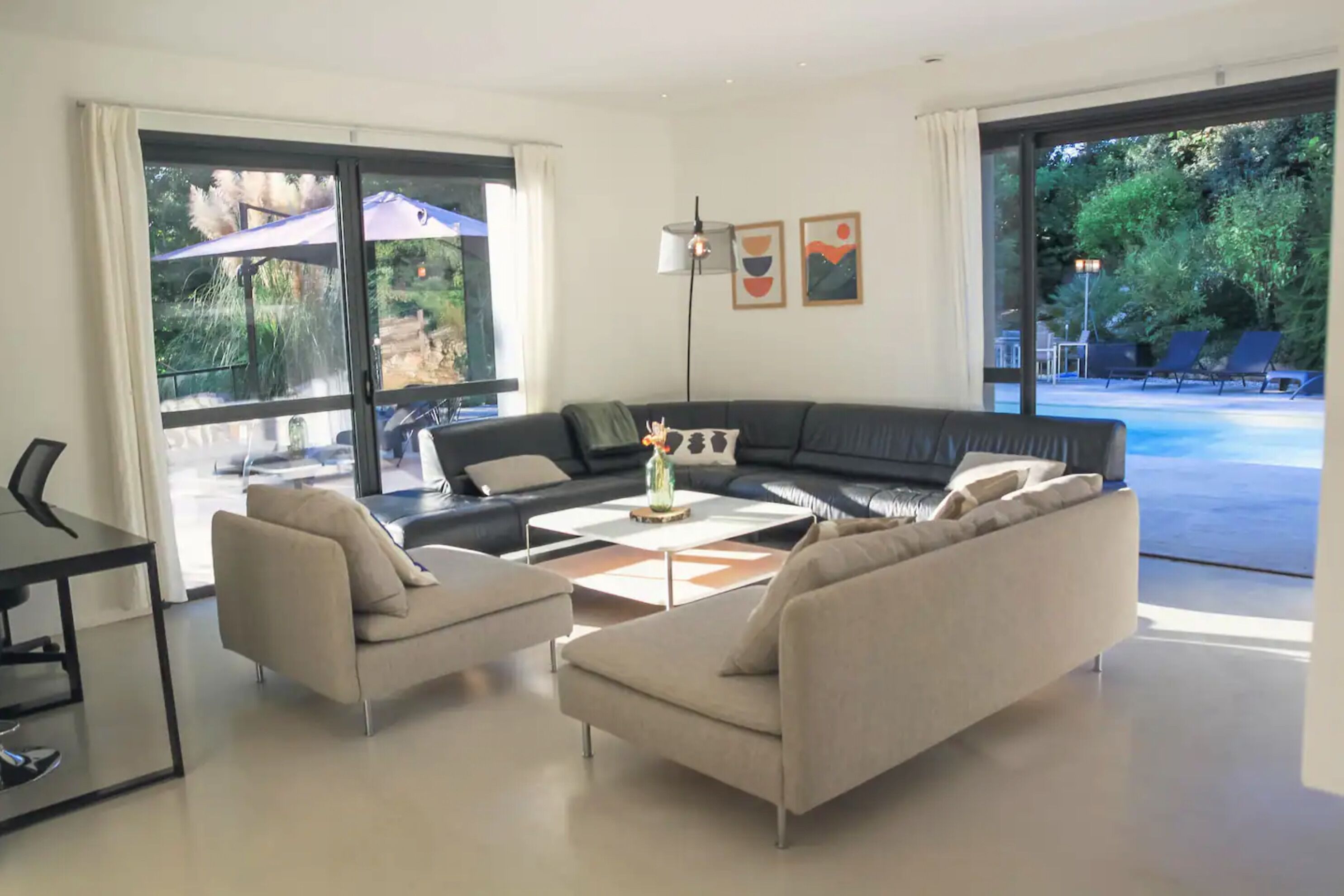 Un salon moderne comprenant un canapé, deux fauteuils, une table basse et une table à manger. De grandes fenêtres offrent une vue sur une piscine et un espace de détente extérieur.