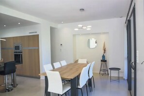 Un espace repas moderne avec une table en bois et des chaises blanches, une cuisine dotée de meubles et d’appareils électroménagers en bois, et une petite table d’appoint sur laquelle est posé un vase.