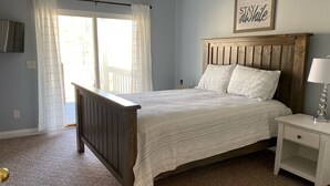 Un dormitorio con cabecera de cama de madera, mesita de noche blanca y vistas al exterior a través de una ventana con cortinas translúcidas.