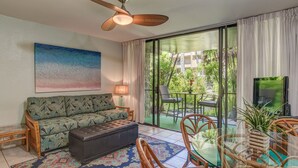 Una sala de estar con un sofá, un reposapiés y una mesa de centro de vidrio. Hay un ventilador de techo, un cuadro y vistas a un jardín a través de una puerta corrediza de cristal.