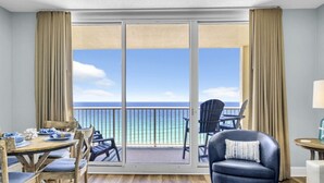 A room with a balcony, a dining table with chairs, a sofa, and a view of the ocean.