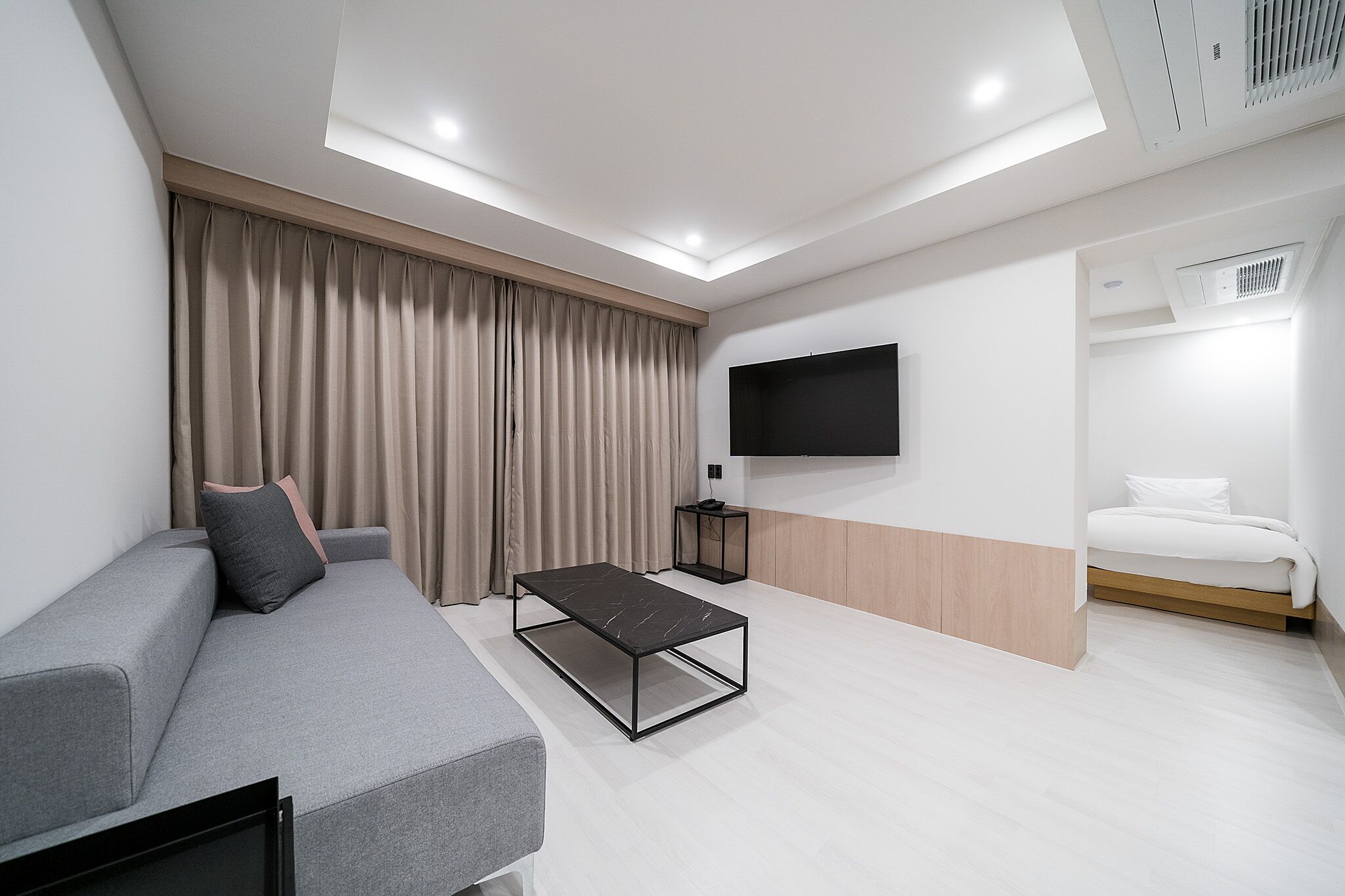 A modern hotel room with a grey sofa, a black coffee table, a bed with white bedding, a TV mounted on the wall, and a ceiling vent.
