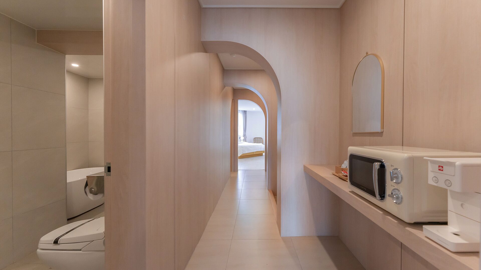 Un pasillo estrecho con un microondas sobre una encimera, que da a un dormitorio con una cama y una silla.