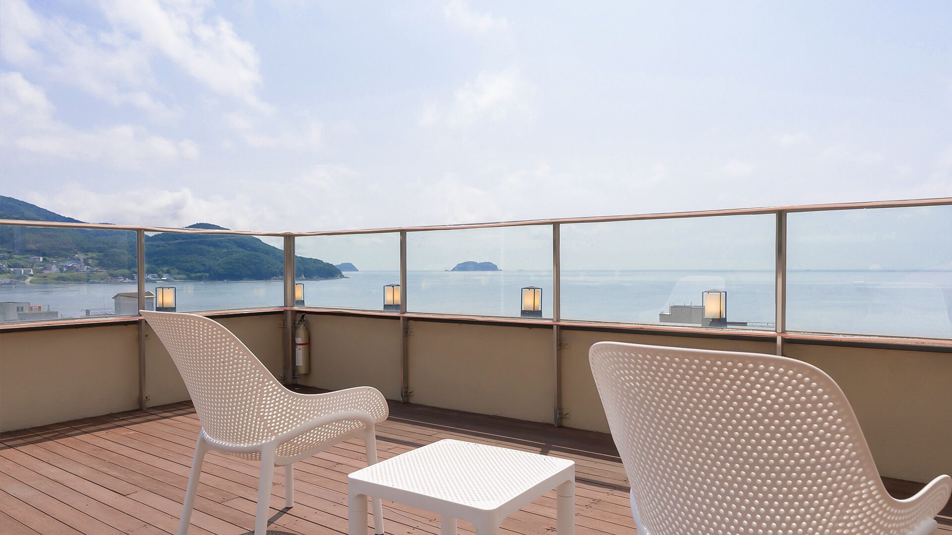 A deck with two white chairs and a small table, overlooking a calm sea with distant islands.