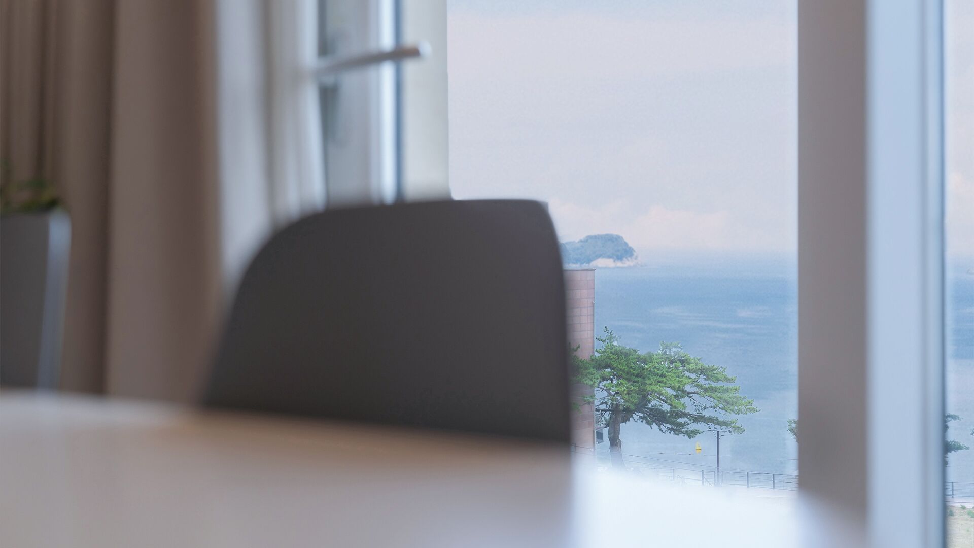 Vista desde una ventana, con una silla al frente, un árbol y un edificio distante sobre el agua.