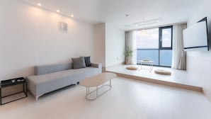 A modern living room with a sofa, a coffee table, and a small side table. There is a large window with a view of the sea. - H.Avenue (Geoje)