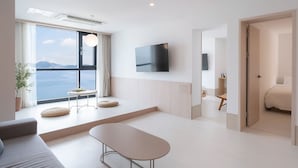A modern hotel room with a large window offering a view of the sea, a flat-screen TV mounted on the wall, a sofa, a small table with cushions, and a bench. - H.Avenue (Geoje)