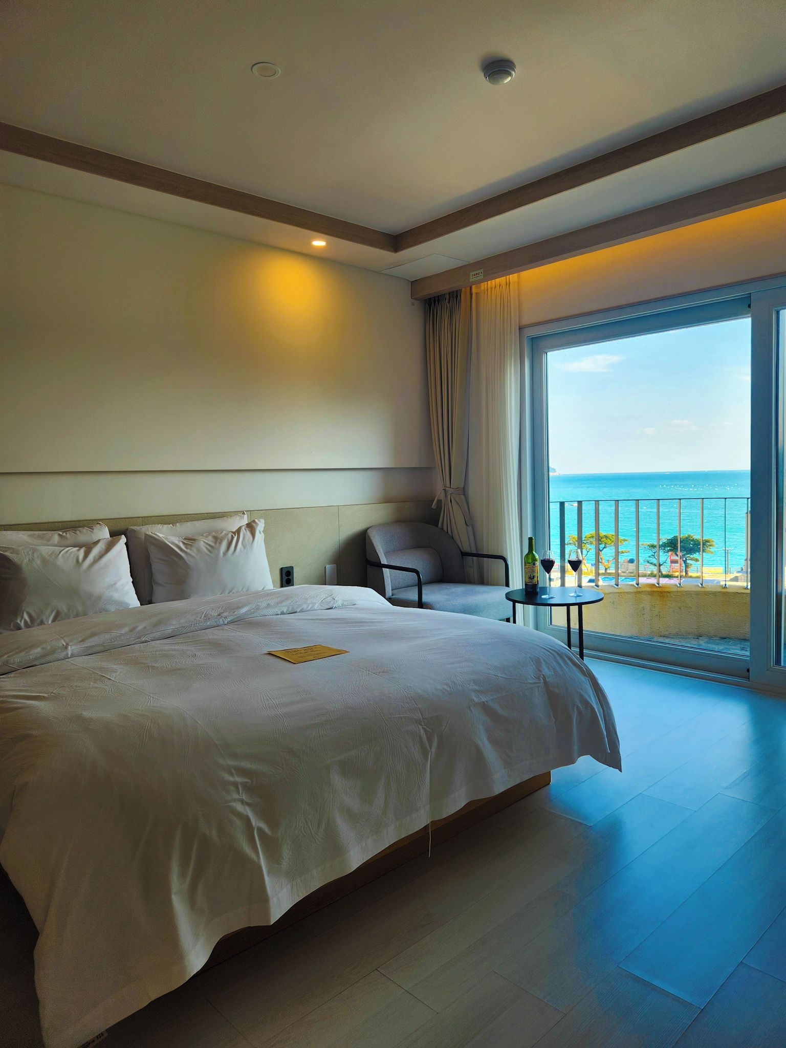 A hotel room with a large bed, a chair, a small table with a bottle and glass, and a balcony with a view of the sea.