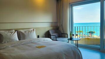 A hotel room with a large bed, a chair, a small table with a bottle and glass, and a balcony with a view of the sea.