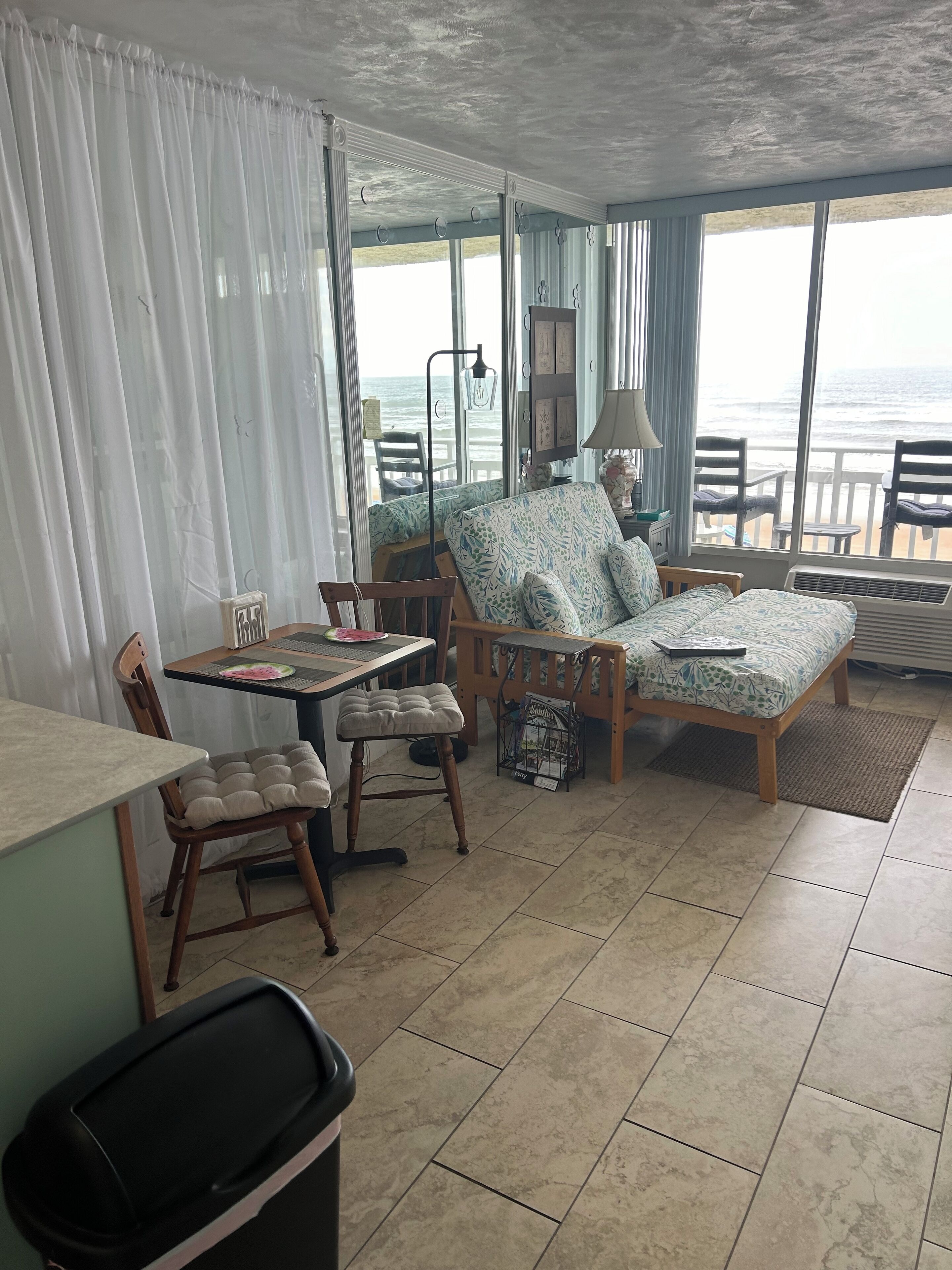 A room with a bed, a desk, and a chair. There is a view of the ocean through large windows.