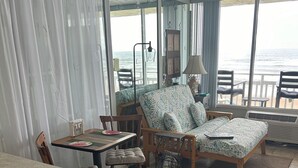 A room with a bed, a desk, and a chair. There is a view of the ocean through large windows.