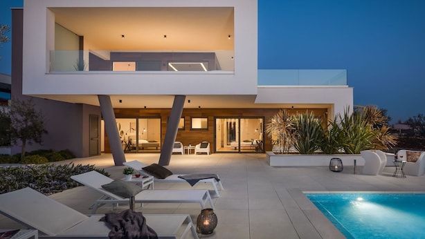 Een modern huis met twee verdiepingen, een zwembad, lounge fauteuils en een goed verlicht interieur.