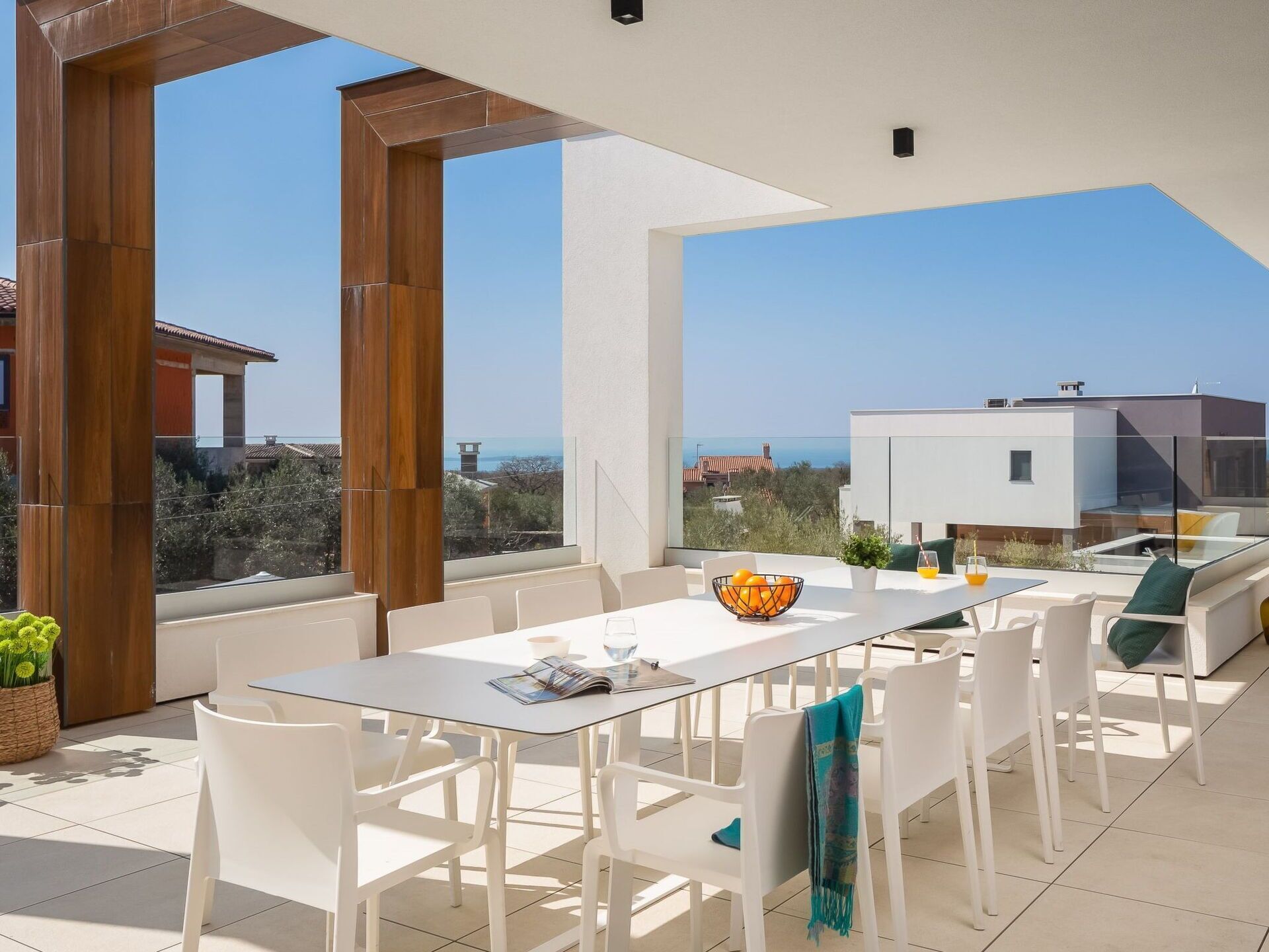 A modern outdoor dining area with a glass table and white chairs, overlooking a scenic view of the sea and buildings.