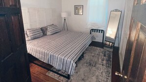 Un dormitorio con cama, lámpara de noche, ventana con persianas, espejo y puerta.