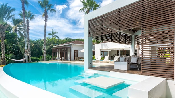Un área de piscina moderna con agua azul cristalina, una pérgola de madera y sillones de descanso.