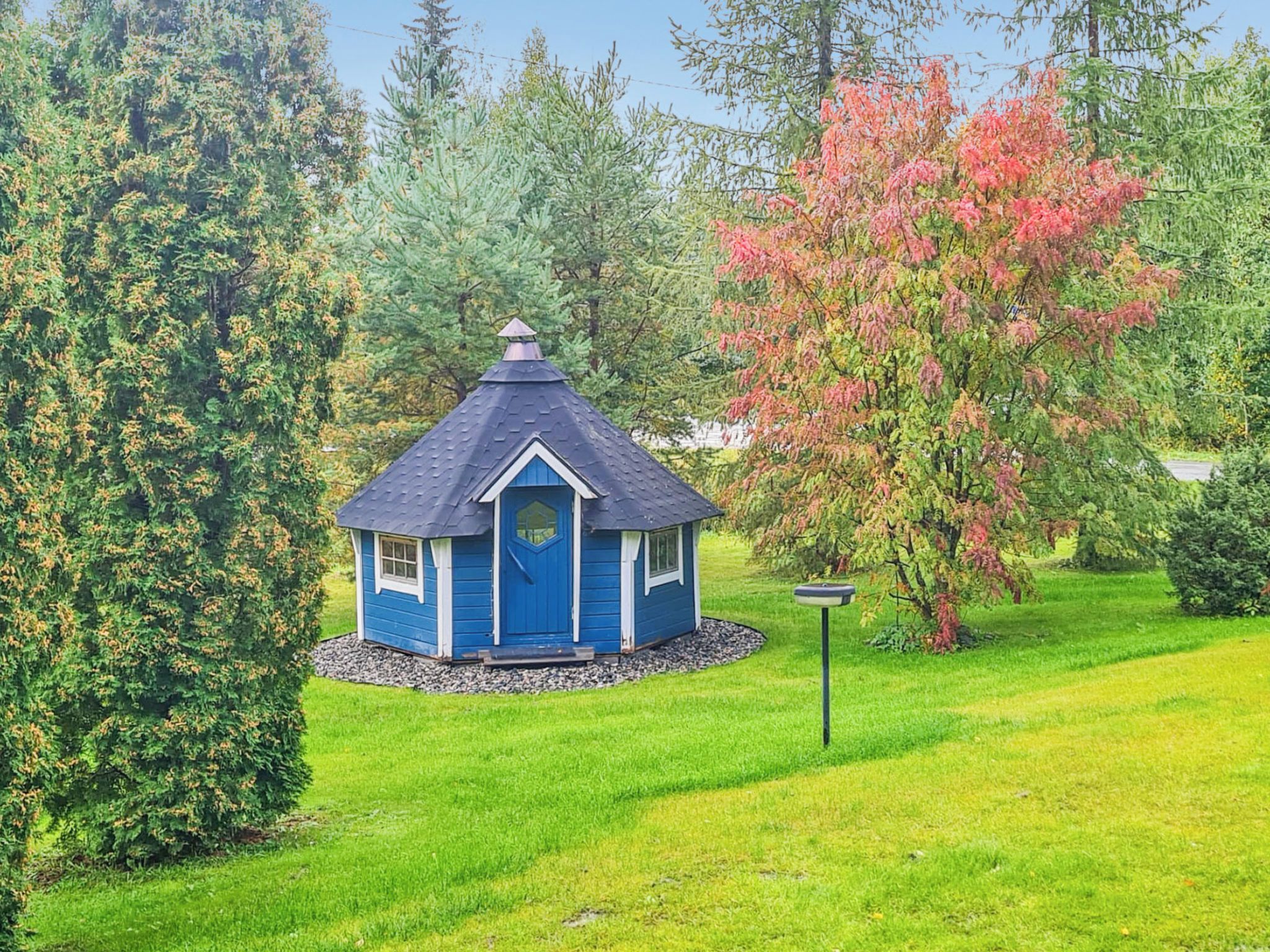 A small blue gazebo with a peaked roof surrounded by lush greenery and trees.